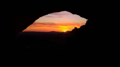 A stunning desert sunset is captured through a natural rock arch, showcasing a dramatic chiaroscuro effect.  The image featur