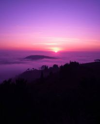 This image captures a mystical sunset over the misty, rolling hills of Tuscany, emanating an ethereal vibe. The scene is char