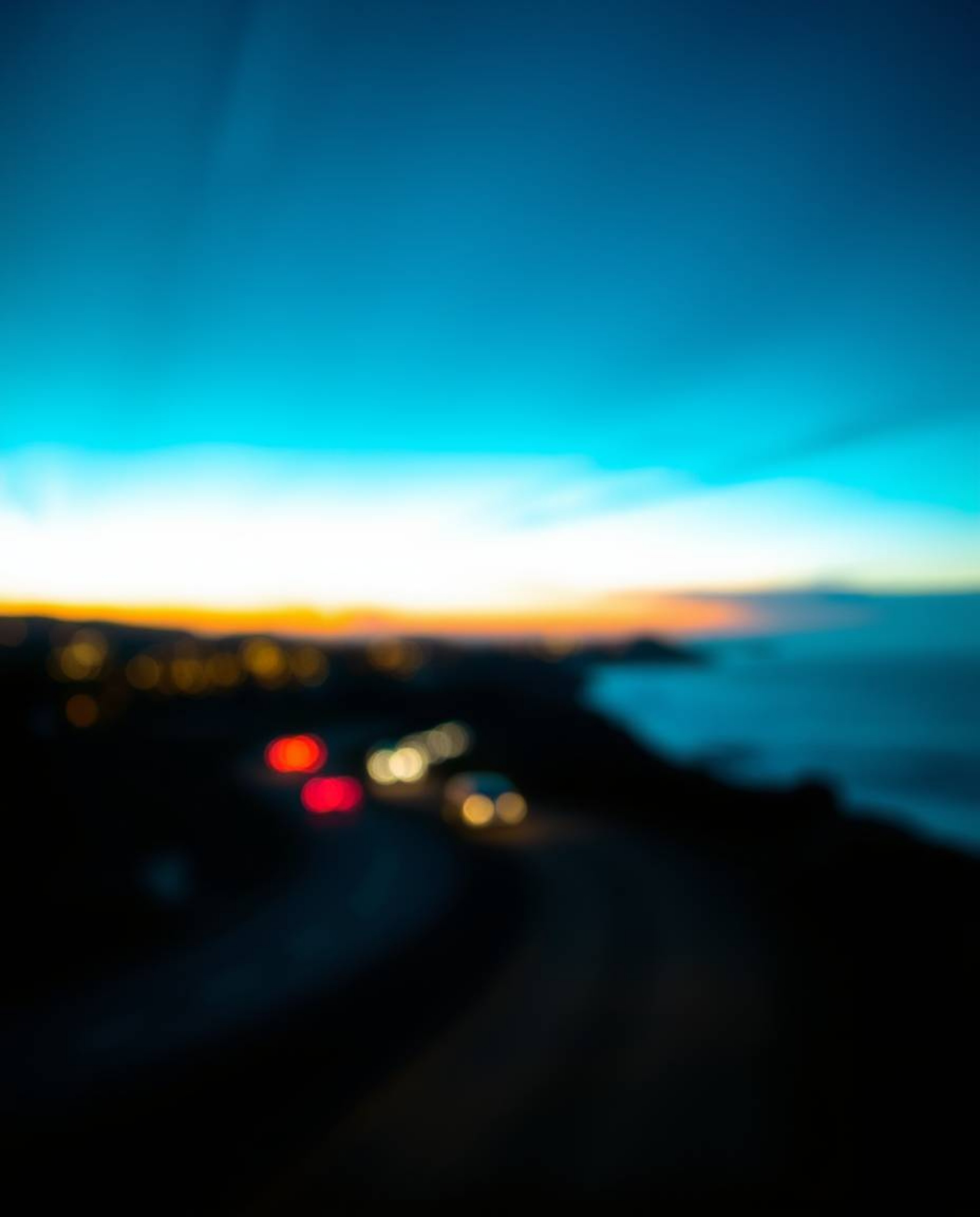 This image captures a rich, deep teal sunset sky over a winding coastal highway, distinguished by a stunning blurred bokeh ef