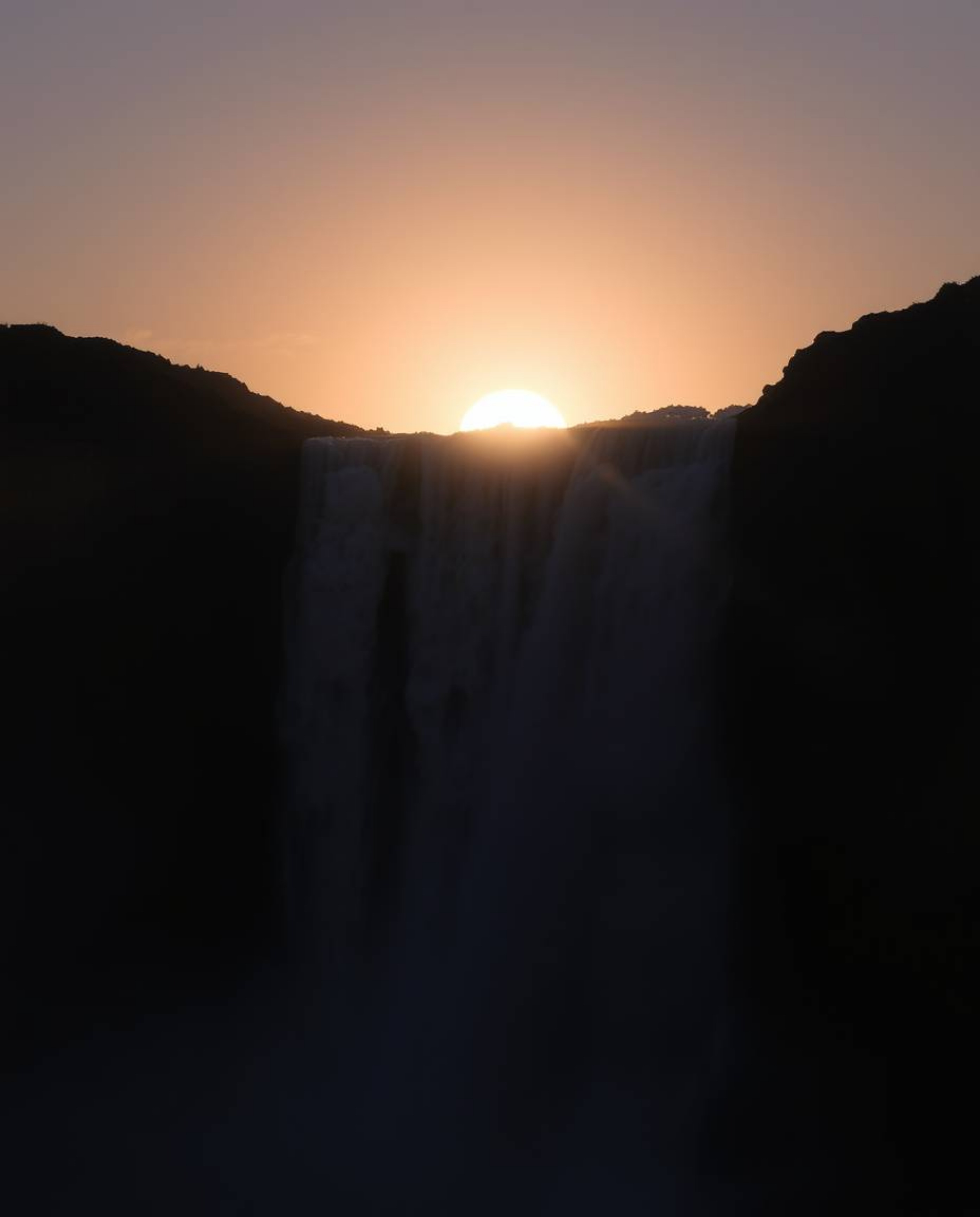 This is a close-up photograph of a sun setting behind a dramatic waterfall. The image is characterized by its use of chiarosc