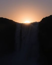 This is a close-up photograph of a sun setting behind a dramatic waterfall. The image is characterized by its use of chiarosc