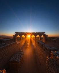 A high-angle perspective photograph captures ancient stone ruins at sunset. The scene features volumetric lighting with visib