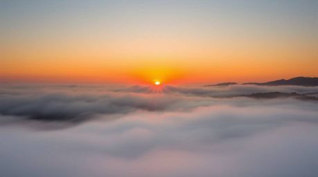 This image captures the ethereal beauty of fog-covered hills bathed in the warm light of a setting sun. The thick fog creates