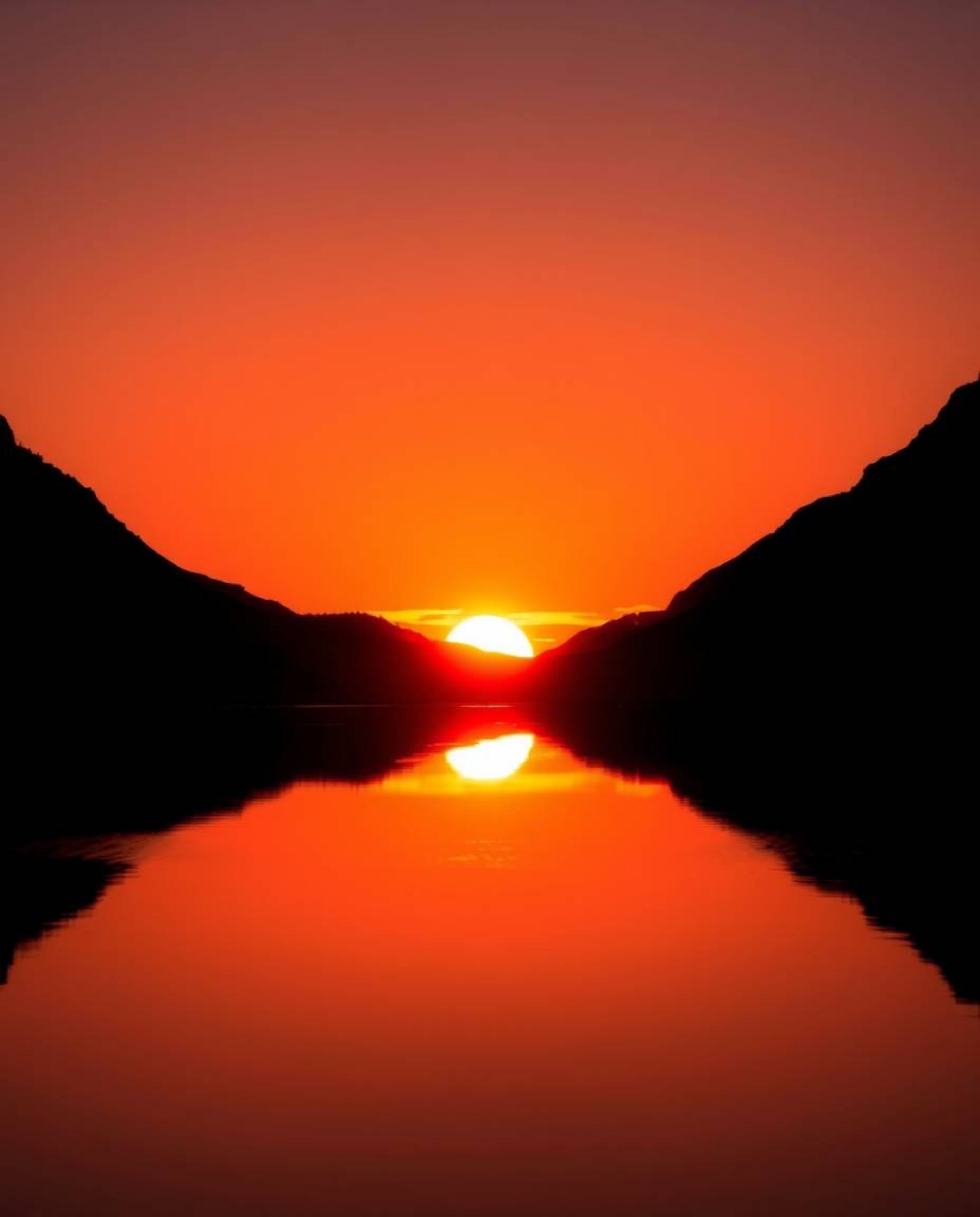 A silhouetted image captures a sunset reflected in a glassy fjord. The scene is defined by warm backlighting, with the light 