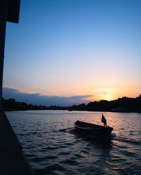 A candid street photograph captures a boat drifting on a lake at sunset, uniquely framed by a visible urban environment. The 