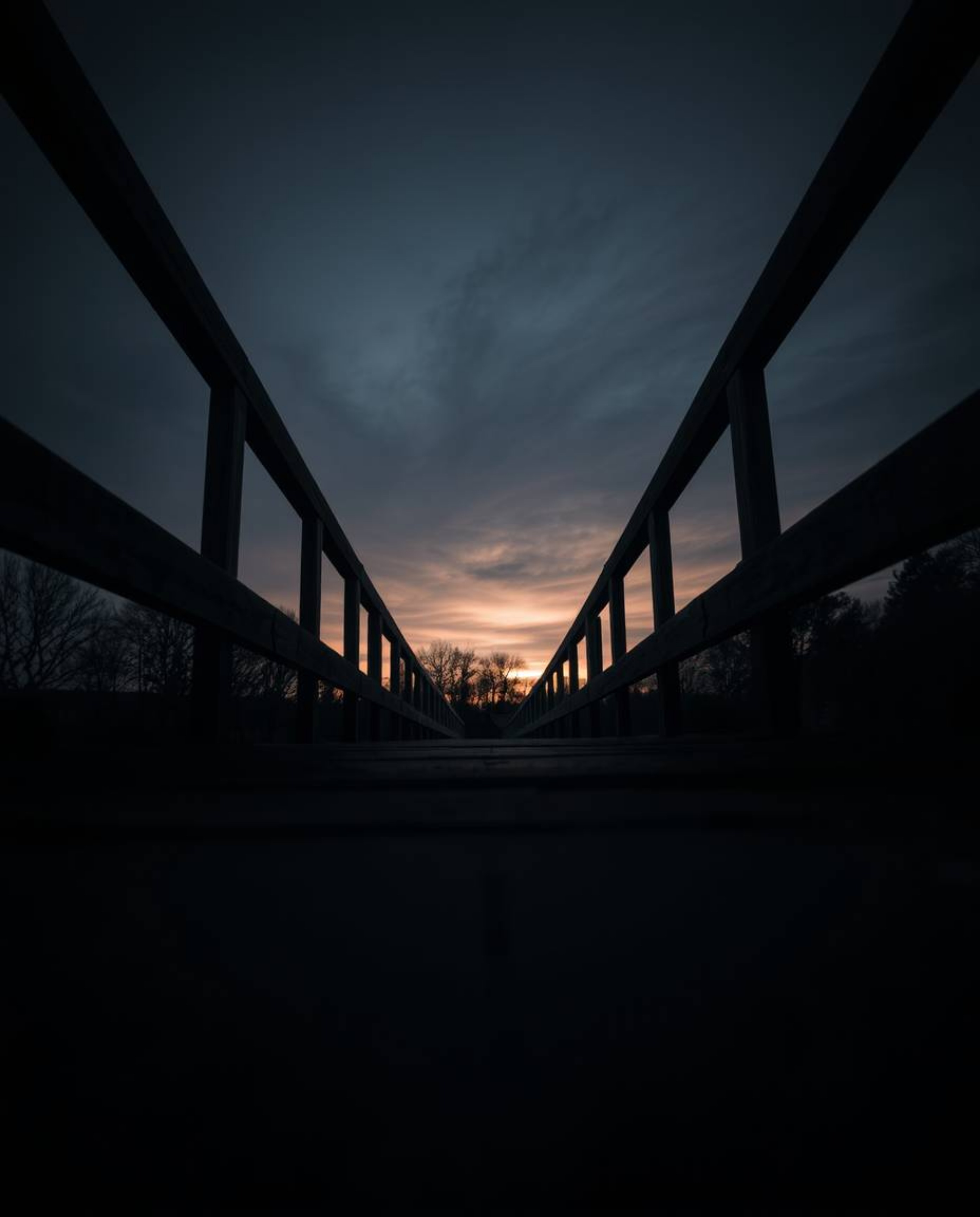 A low-angle perspective of an old wooden bridge over a river at sunset. This photograph is presented in a moody style with lo