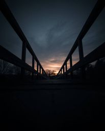 A low-angle perspective of an old wooden bridge over a river at sunset. This photograph is presented in a moody style with lo