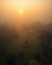 A high-angle, fine art photograph depicting a serene Zen garden at sunset shrouded in thick fog, exhibiting warm, soft tones 