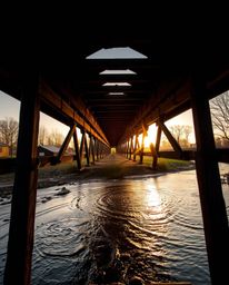 A dramatic scene of an old wooden bridge over a river at sunset, captured from a low-angle perspective looking up. The compos