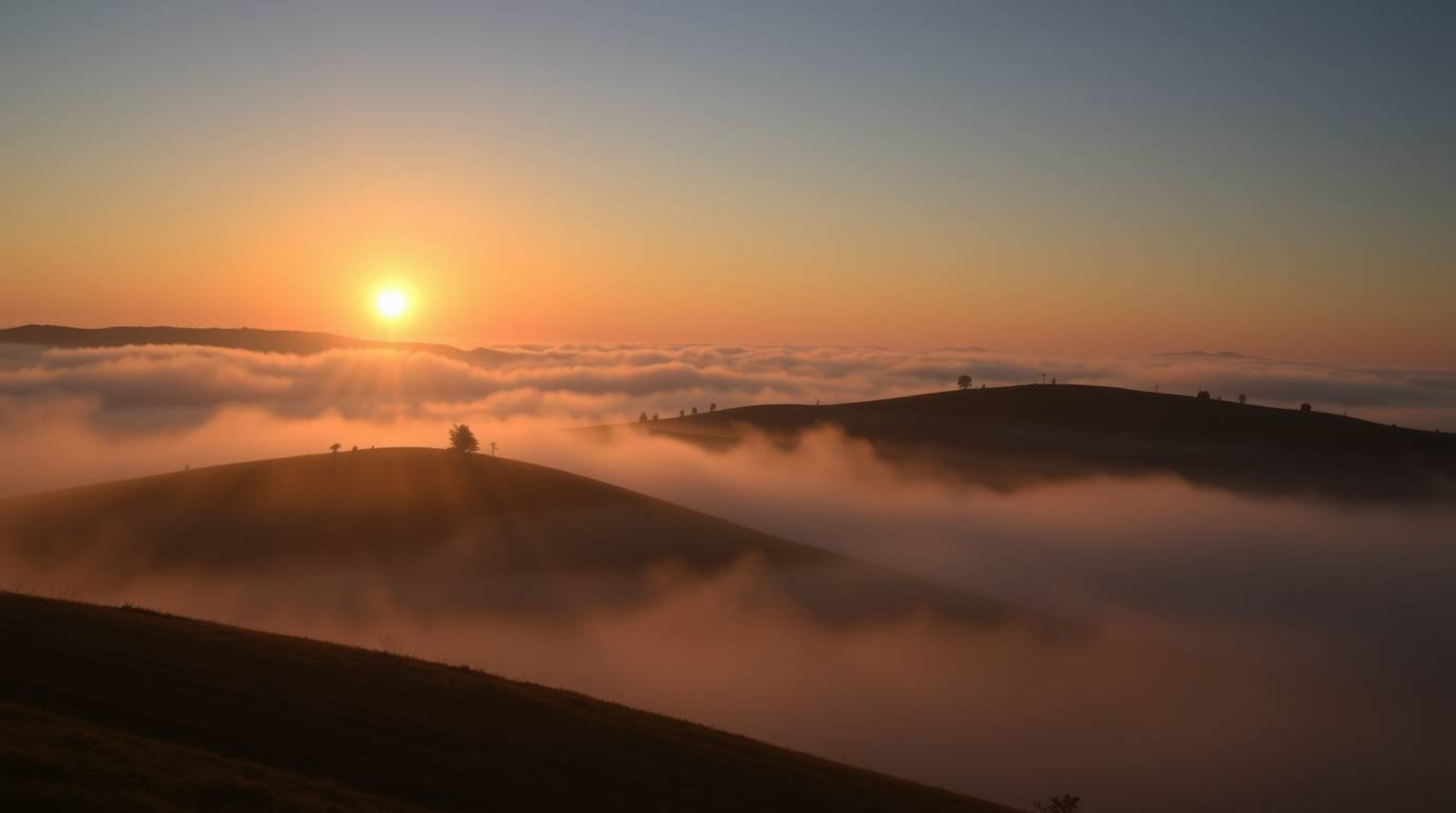 Capture the serene beauty of fog-covered hills bathed in the golden light of a setting sun. This image boasts a graceful eleg