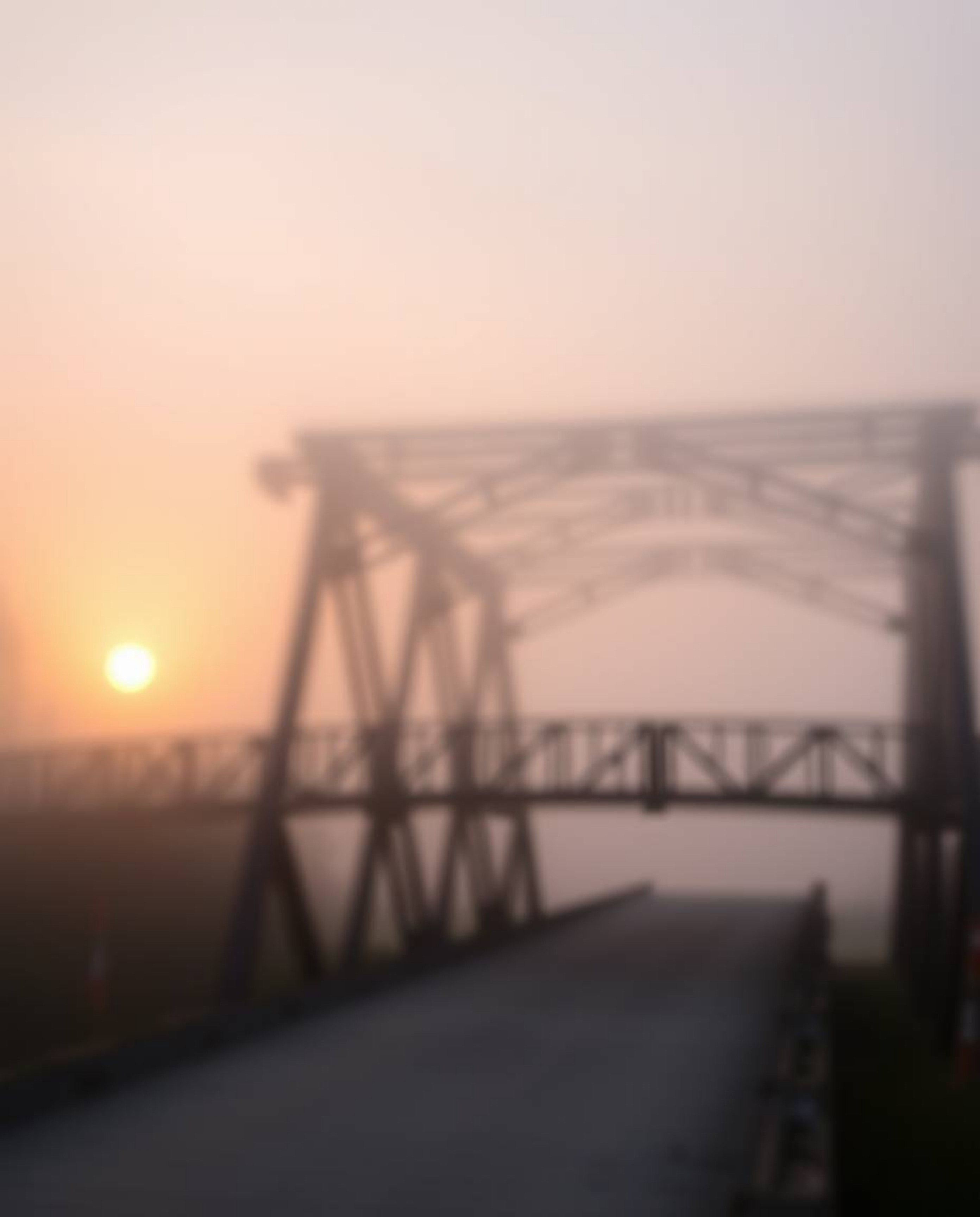 A photograph captures an old wooden bridge over a river at sunset, enveloped in thick fog, which creates a scene of low visib