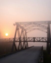 A photograph captures an old wooden bridge over a river at sunset, enveloped in thick fog, which creates a scene of low visib