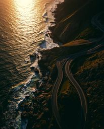 An overhead bird’s-eye view captures a winding coastal highway at sunset, presented from a dramatic top-down perspective. The