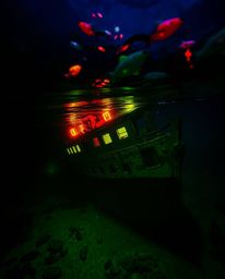 This candid street photography captures a shipwreck illuminated deep underwater, glowing with bioluminescent reds, oranges, a