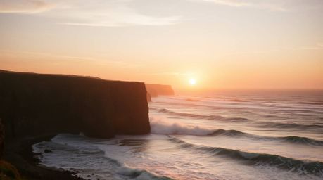 Experience the warm embrace of autumnal tones as waves crash dramatically against cliffs beneath a blazing sunset sky. The sc