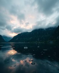 A striking photograph taken from a low-angle perspective of a glassy fjord reflecting a sunset in a wet, rainy scene. The ima