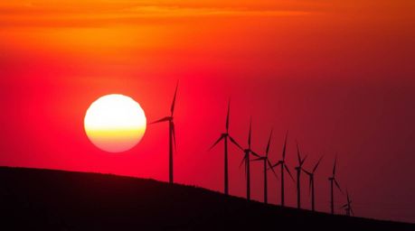 A stunning close-up captures a warm sunset painting the sky in rich reds, oranges, and yellows behind a row of wind turbines 