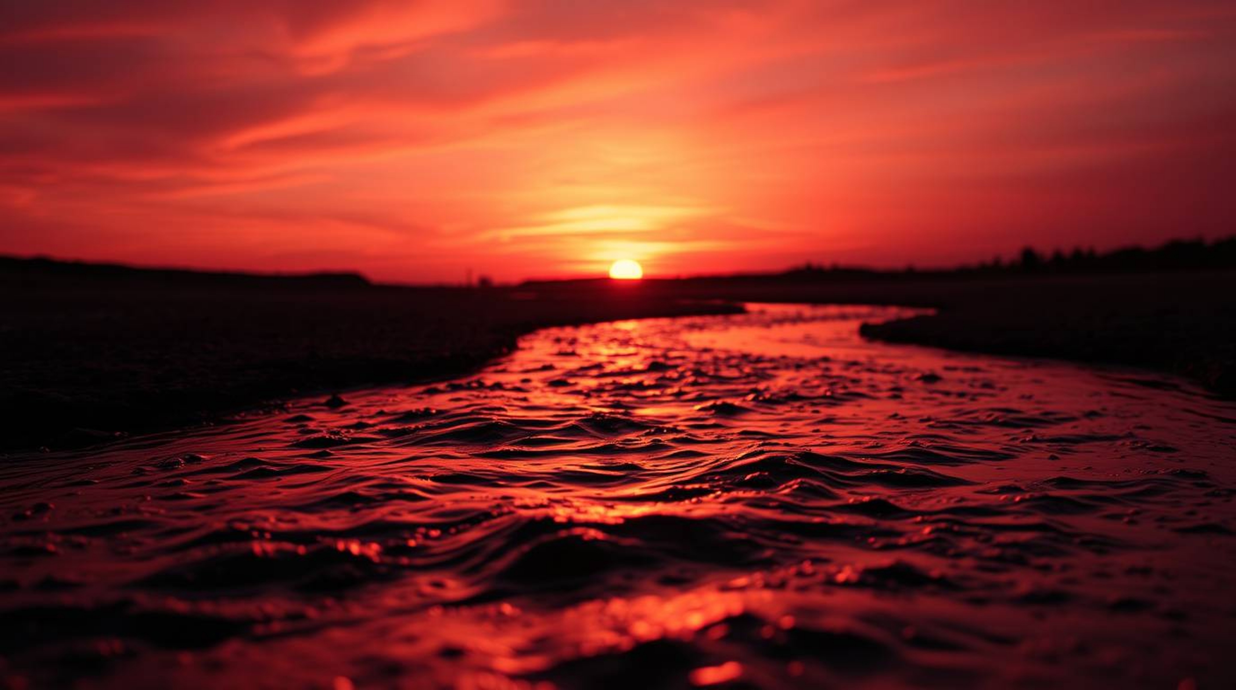A stunning macro close-up photograph showcasing a winding river illuminated by a dramatic orange-pink sky. The silhouetted sc