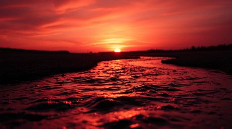 A stunning macro close-up photograph showcasing a winding river illuminated by a dramatic orange-pink sky. The silhouetted sc