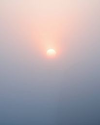 Thick fog and soft pastel colors characterize this scene of a sun setting behind a dramatic waterfall. The overall effect is 