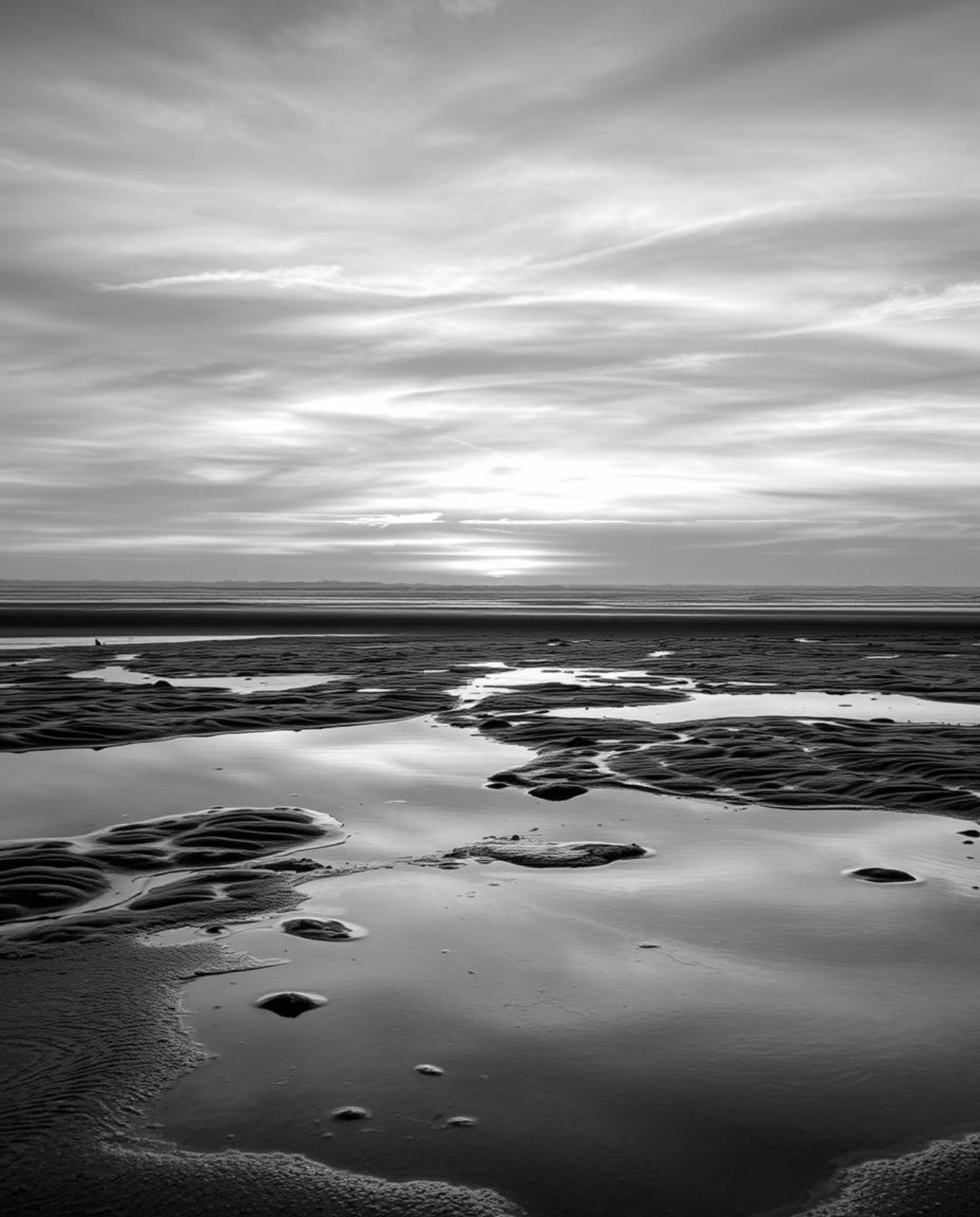 A classic black and white photograph captures a wet rainy scene, showcasing a sunset sky mirrored in tidal pools on the beach