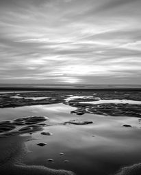 A classic black and white photograph captures a wet rainy scene, showcasing a sunset sky mirrored in tidal pools on the beach