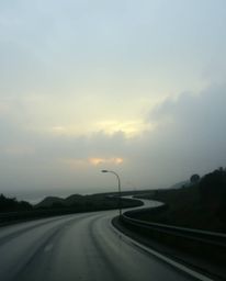 This is a soft, dreamy depiction of a wet, rainy scene featuring a sunset sky over a winding coastal highway. The atmosphere 