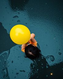 A captivating overhead shot of a child with dark hair holding a bright yellow balloon on a rainy day. Reflective puddles and 