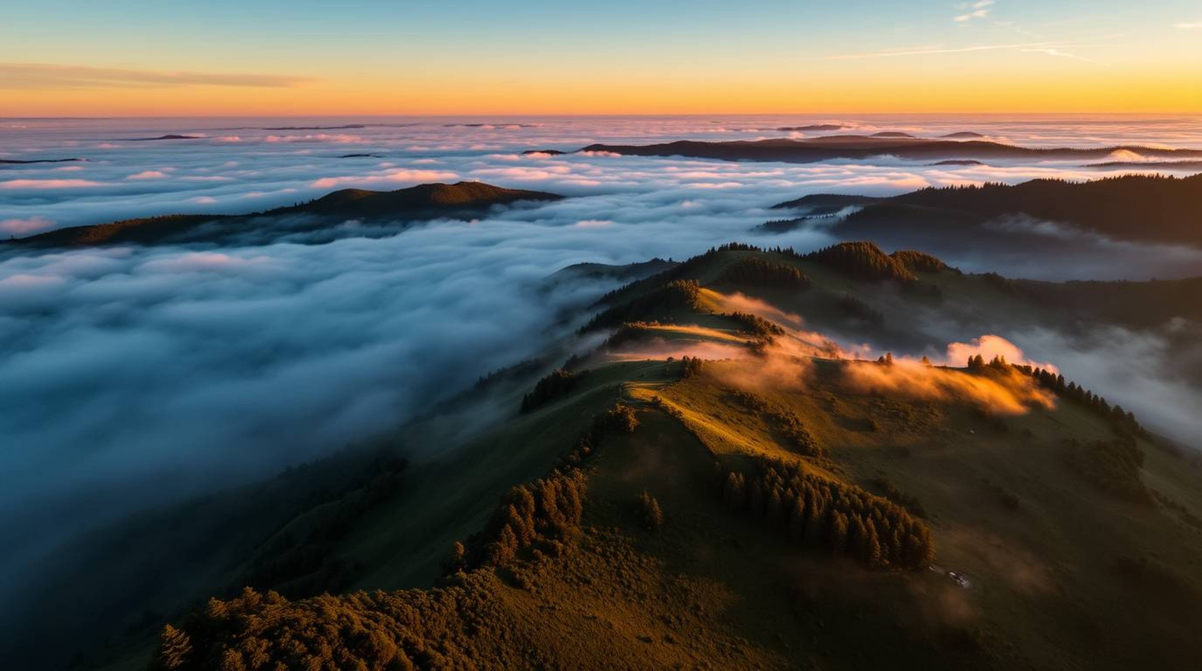 Experience the serene beauty of fog-covered hills bathed in the warm glow of a setting sun from a unique overhead bird's-eye 