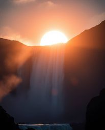 This is a cinematic and epic scene featuring a massive waterfall silhouetted against the setting sun. The composition is enha
