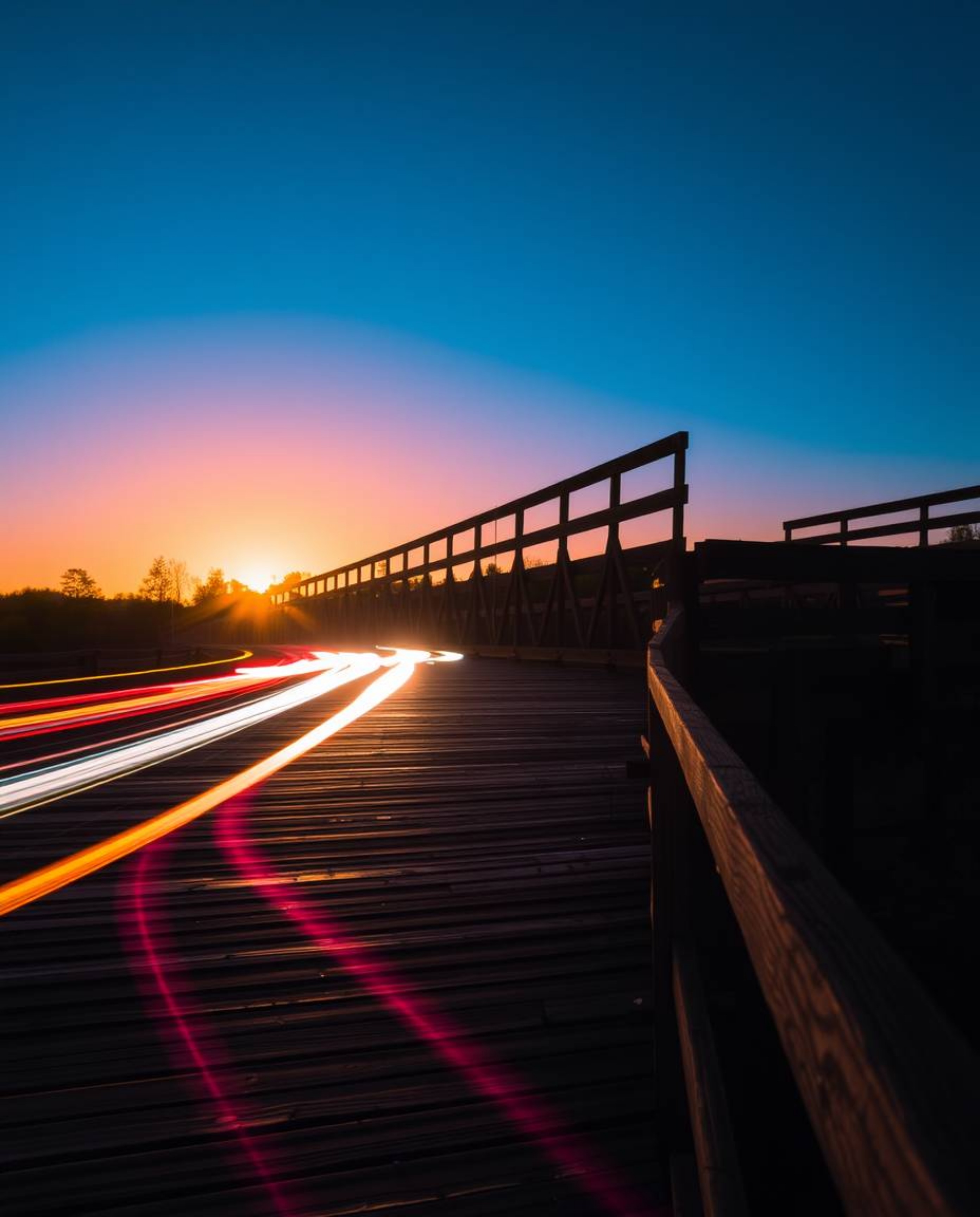 This is a long-exposure photograph of an old wooden bridge over a river at sunset. The image features bright, vibrant colors,