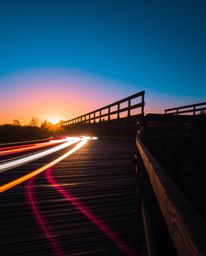 This is a long-exposure photograph of an old wooden bridge over a river at sunset. The image features bright, vibrant colors,