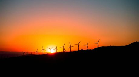 A vibrant sunset paints the sky in complimentary colors, silhouetting a row of wind turbines atop a hill. The backlighting cr