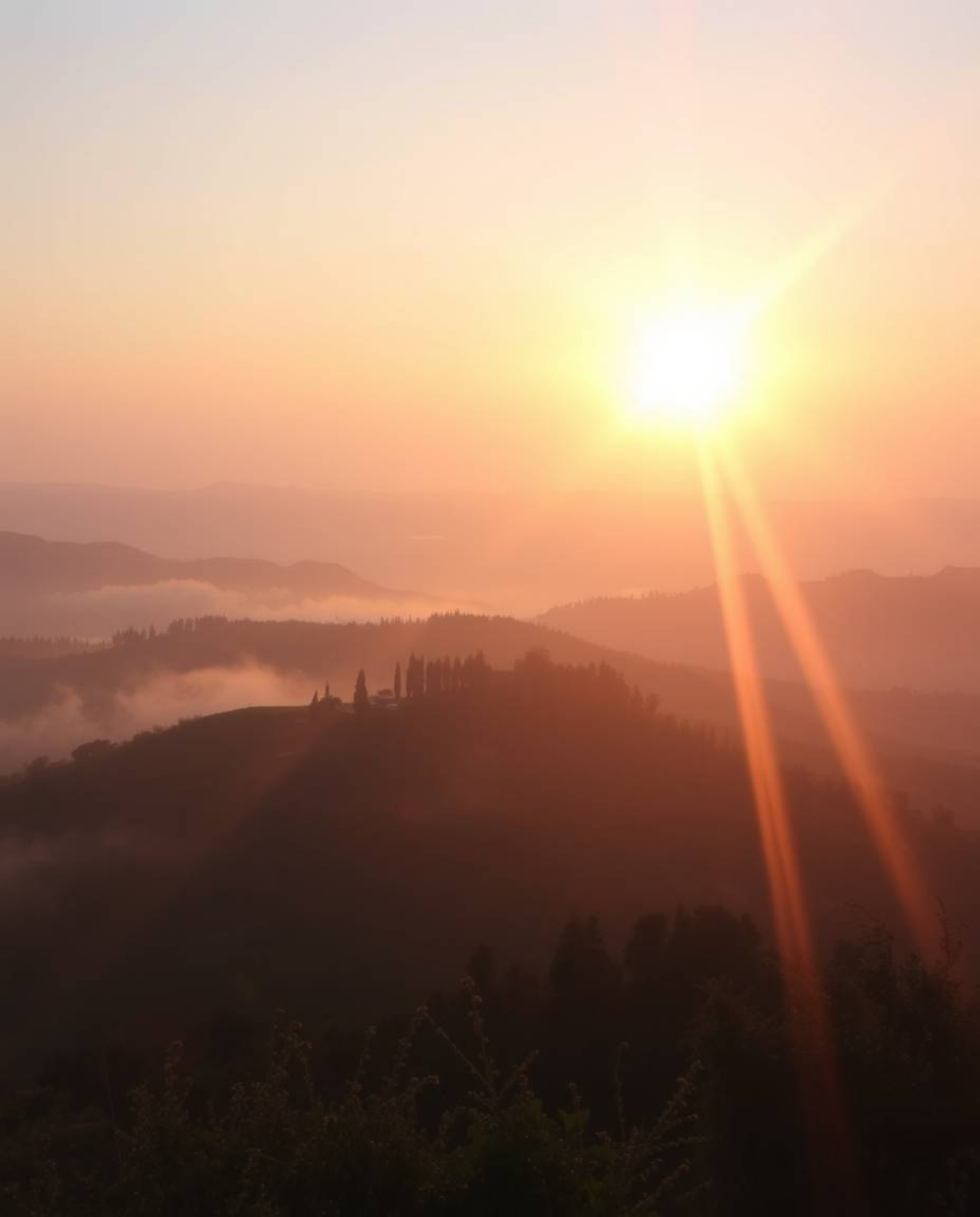 A soft pastel image of a Tuscan sunset over misty hills, viewed from above, featuring bright lens flares, light streaks, and 