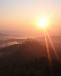 A soft pastel image of a Tuscan sunset over misty hills, viewed from above, featuring bright lens flares, light streaks, and 