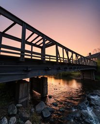 A precise architectural color photograph capturing an old wooden bridge over a river during sunset. This image emphasizes sha