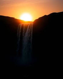 This is a cinematic film still capturing a dramatic waterfall against a setting sun. The entire scene is rendered in intense 