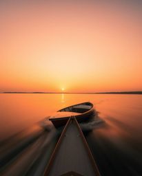 A long exposure photograph captures a graceful and elegant boat drifting on a lake at sunset, its refined features softened b