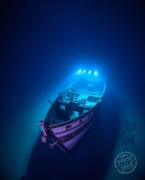 This captivating photograph captures a shipwreck illuminated deep underwater, utilizing a cool color palette of blues, greens