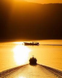Chiaroscuro lighting dramatically illuminates a boat drifting on a lake at sunset, creating a powerful and moody scene. The t