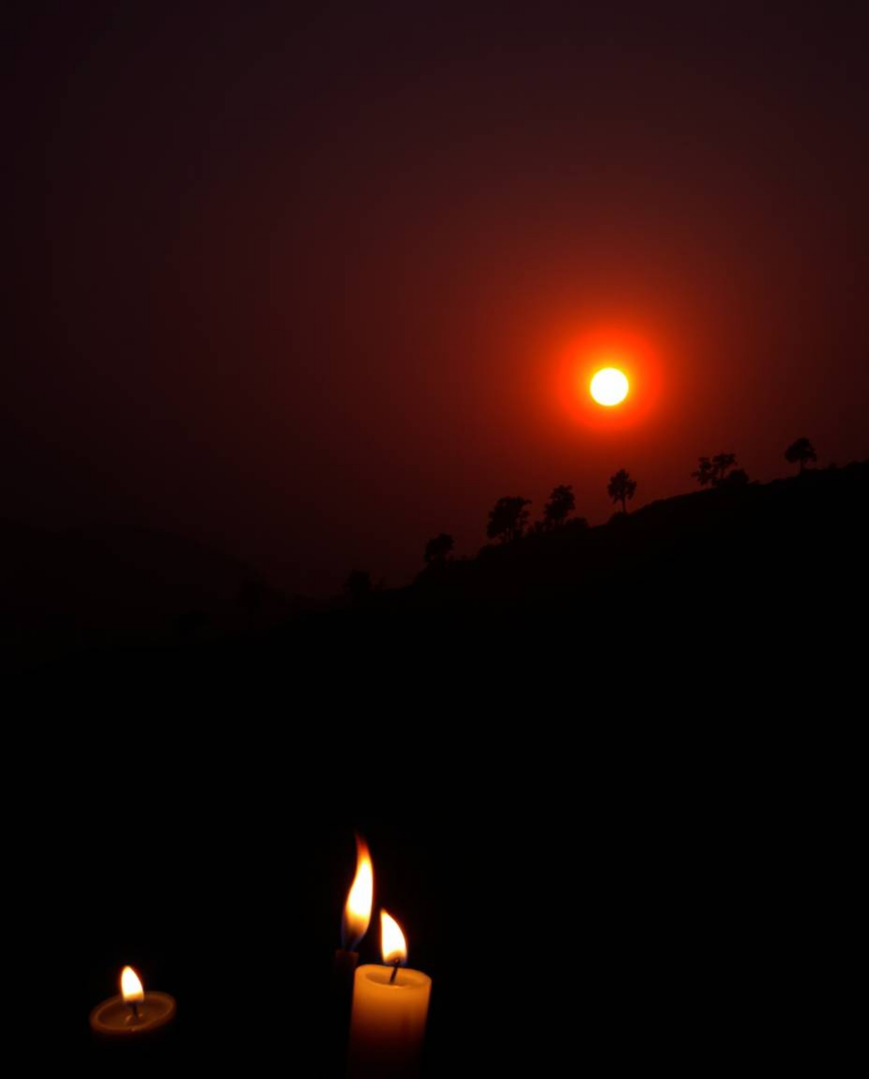A moody, low-contrast photograph depicting a sunset over terraced tea plantations. The scene is illuminated by the warm glow 