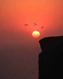 A warm, slightly dark, and soft professional fine art photograph captures a graceful and elegant sunset over a coastal cliff.