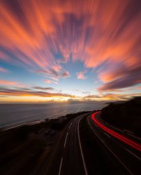This dynamic light art photograph captures a sunset sky over a winding coastal highway. The scene is defined by its artistic 