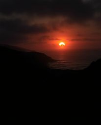 The sunset over a winding coastal highway is depicted with an eerie atmosphere, emphasized by strong hard light and harsh sha