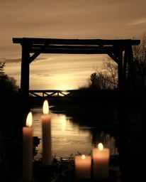 A classic black and white photograph of an old wooden bridge over a river at sunset. The scene is uniquely illuminated by war
