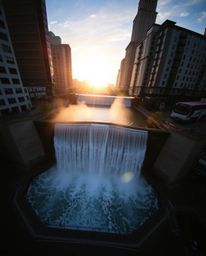 An overhead bird’s-eye view candid street photograph captures a sun setting behind a dramatic waterfall. The image blends urb