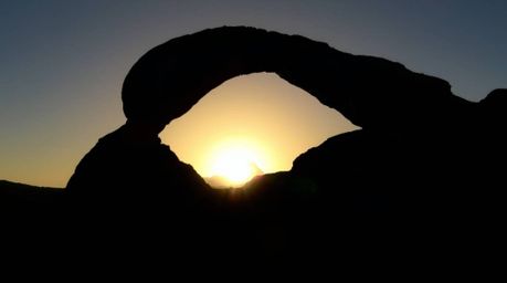 A stunning 16:9 landscape photograph showcasing a silhouetted scene of a split sunset filtering through a natural desert rock