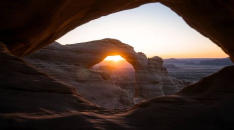 A stunning long-exposure photograph capturing a desert sunset framed by a natural rock arch. The image features gentle, soft 