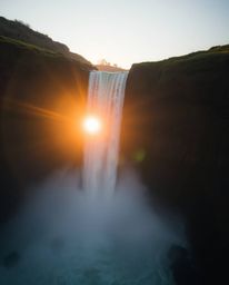 A gentle soft light illuminates an overhead bird’s eye view of a sun setting behind a dramatic waterfall. This top-down persp