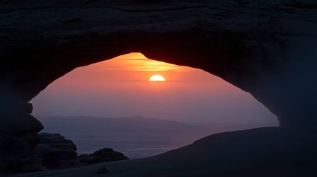 A captivating photograph showcases a muted sunset viewed through a natural rock arch in a desert setting.  Thick fog dramatic
